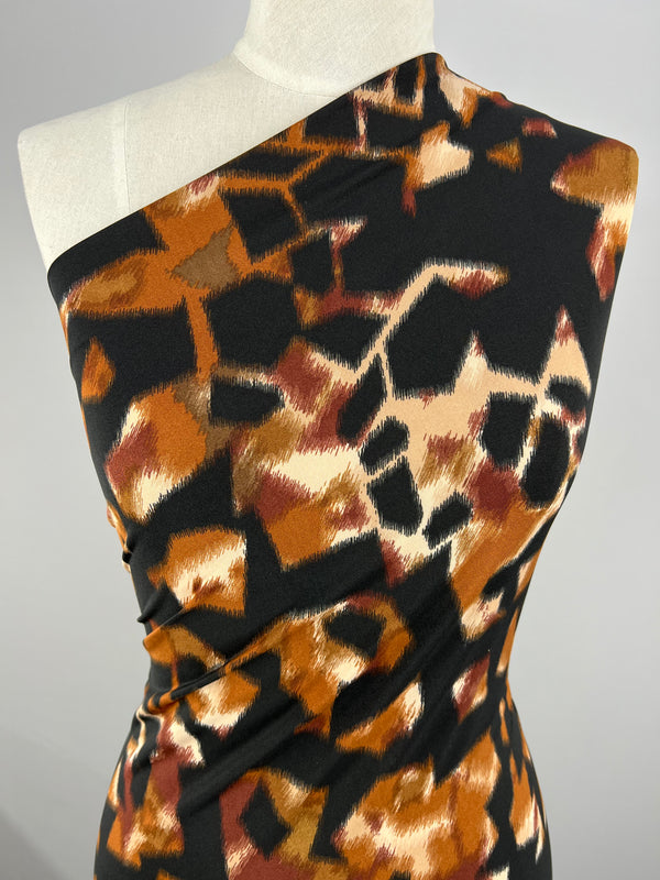 A dress form is draped with a one-shouldered garment made from Super Cheap Fabrics' Printed Lycra - Blurred Blocks, a medium weight polyester spandex blend featuring an abstract pattern in shades of black, brown, orange, and beige. The design appears to mimic brush strokes or scattered geometric shapes inspired by untamed wilderness.