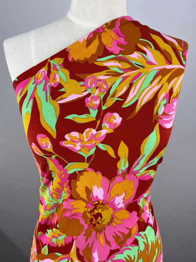 A mannequin wearing a vibrant, off-the-shoulder dress crafted from medium weight polyester fabric with a bold floral pattern in shades of red, pink, yellow, green, and orange. The background is neutral gray, highlighting the colorful design of this versatile piece of multi-use clothing made from Printed Lycra - Ferngully - 150cm by Super Cheap Fabrics.