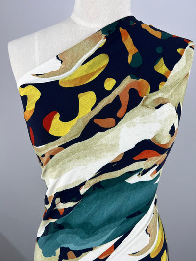 A close-up of a mannequin dressed in a colorful one-shoulder dress made from Super Cheap Fabrics' Printed Lycra - Melt - 150cm. The dress features an abstract pattern with vivid splashes of yellow, green, orange, and black set against a white background, complemented by teal backing for added depth.