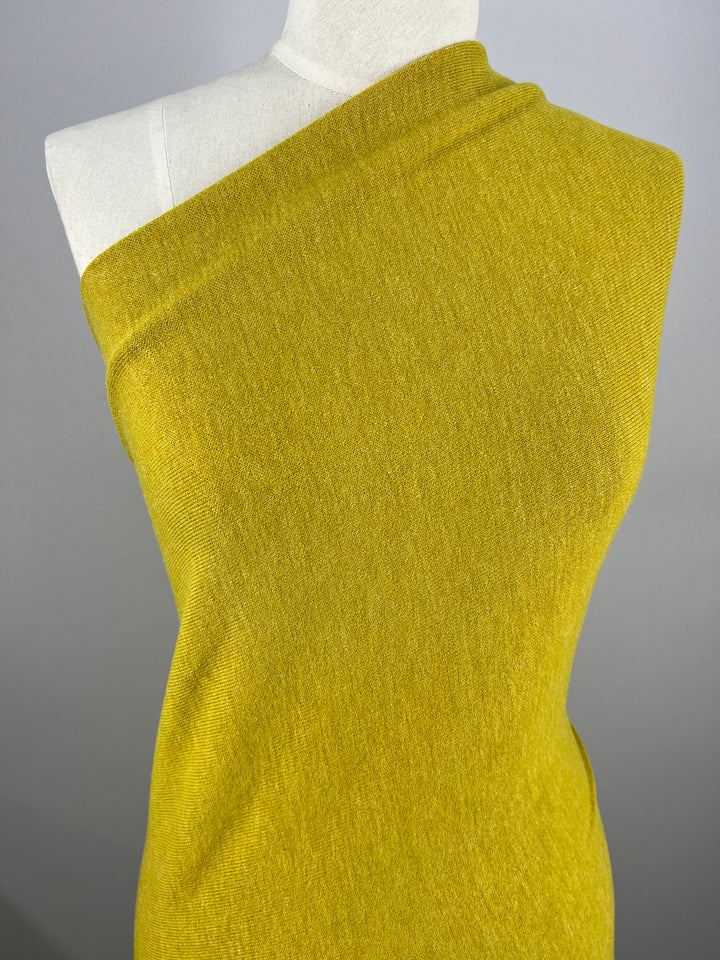 A close-up of a mannequin dressed in a one-shoulder, mustard yellow garment made from Super Cheap Fabrics' Textured Knit - Gold Flake - 145cm. The medium-weight fabric appears soft and slightly textured with hints of gold flake. The background is neutral, making the vibrant color of the dress stand out.