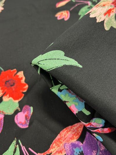 A close-up of *Super Cheap Fabrics' Cotton Sateen - Animate Flora - 125cm* featuring a vibrant floral design on a black background. The multi-color fabric showcases flowers in red, pink, and purple hues with green leaves. It has been partially folded over on the right side of the image, revealing the back of the lightweight fabric.