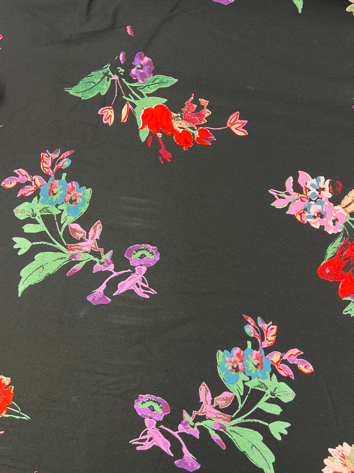 A close-up of a dark cotton fabric with a colorful floral pattern. The flowers include vivid shades of red, pink, purple, and blue with green leaves. The intricate design stands out against the dark background, creating a vibrant and lively visual. This is Cotton Sateen - Animate Flora - 125cm by Super Cheap Fabrics.