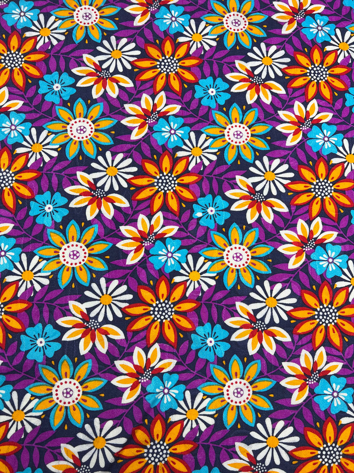 A colorful, lightweight fabric pattern featuring a dense, repeating arrangement of flowers in orange, yellow, blue, and white with red centers on a dark purple background. The multi-colour vibrant flowers are intertwined with teal leaves, creating a lively, intricate design perfect for dresses. This is the Printed Rayon - Lolita Evening - 145cm by Super Cheap Fabrics.