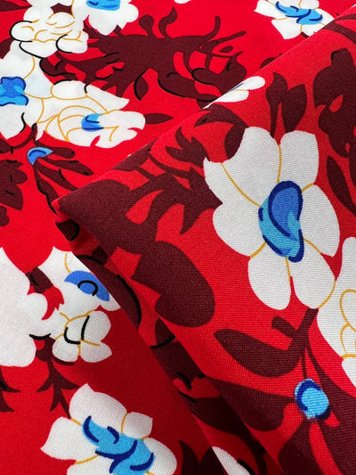 A piece of Printed Rayon - Alice - 150cm from Super Cheap Fabrics with a vibrant red background featuring a floral pattern with white flowers, blue centers, and dark red and gold outlines. This lightweight, multi-colour fabric is folded, showcasing the repeating design.