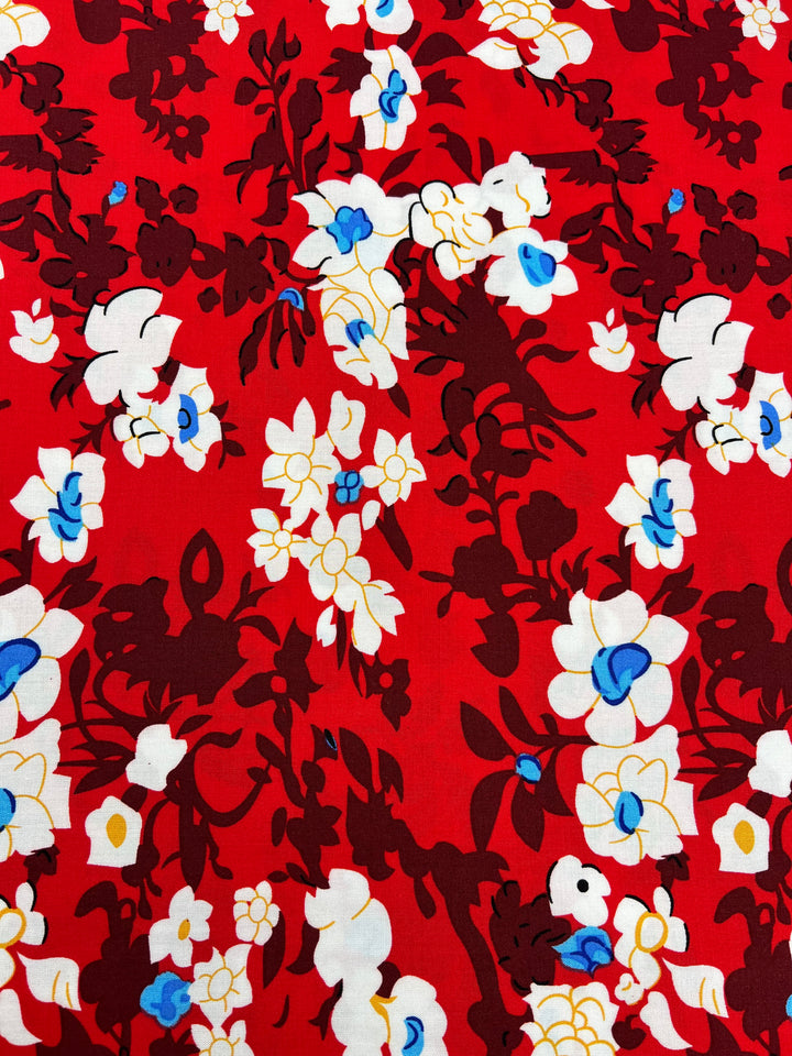 A vibrant red fabric with an intricate floral pattern. The design, crafted from lightweight 100% rayon, features white and yellow flowers with blue centers and dark maroon leaves, creating a bold and eye-catching multi-colour contrast across the fabric. This is the Printed Rayon - Alice - 150cm by Super Cheap Fabrics.