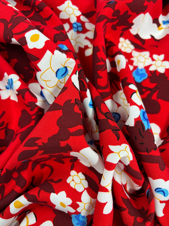 A close-up of a vibrant red fabric with a floral pattern. The design features white, yellow, and blue flowers interspersed with dark red and black abstract shapes, creating a lively and colorful texture. Made from 100% rayon, the light-weight fabric appears soft and slightly draped. This is the Printed Rayon - Alice - 150cm by Super Cheap Fabrics.