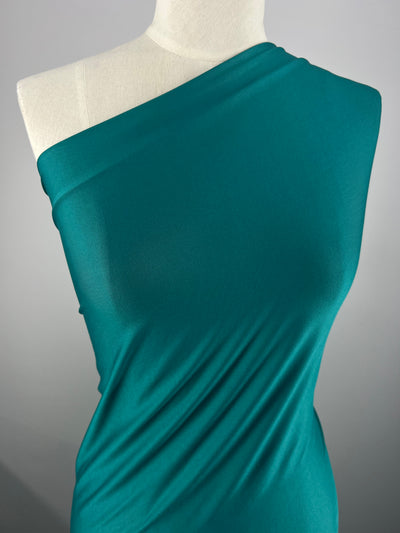A mannequin draped in a teal, one-shoulder Plain Lycra - Tile - 160cm from Super Cheap Fabrics that drapes smoothly across the torso. The versatile fabric has a soft, slightly shiny texture and showcases gentle folds and contours, creating form-fitting garments. The background is a simple, neutral gray.