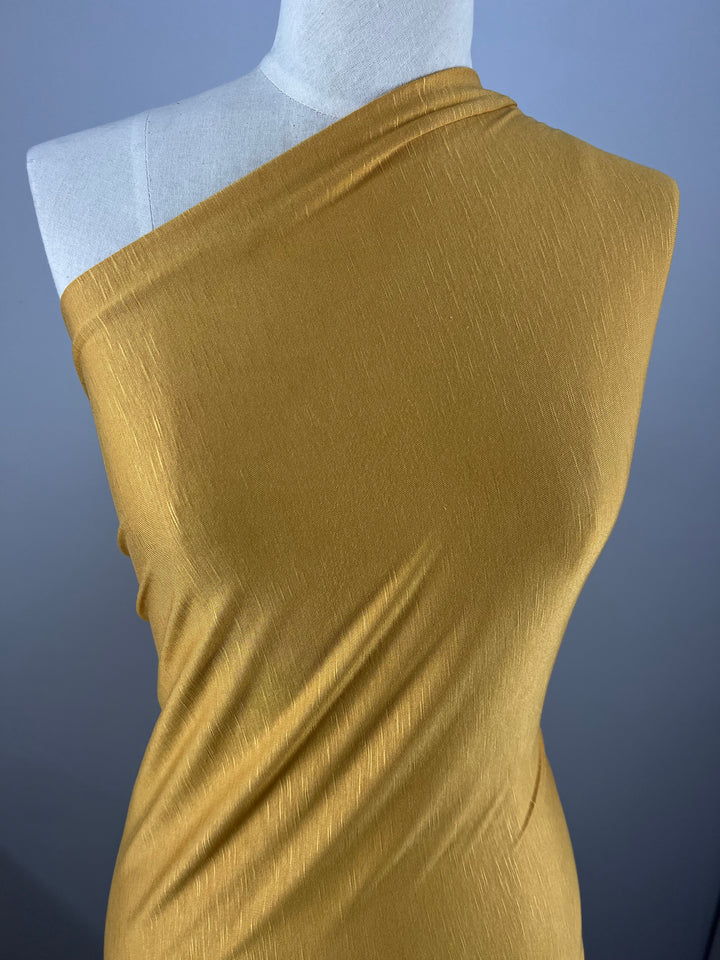 A mannequin is draped in an environmentally responsible, golden-yellow, one-shoulder fabric with a subtle texture. The material appears smooth and slightly shiny, creating gentle folds and curves along the mannequin's form. The background is plain and gray. The fabric used is Bamboo Jersey - Arrowwood - 160cm by Super Cheap Fabrics.