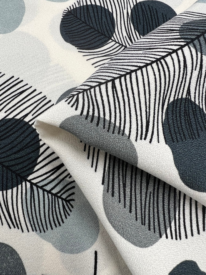 Close-up of the lightweight Printed Crepe - Black Dotty Leafs fabric from Super Cheap Fabrics, showcasing its modern, abstract design with black line-drawn leaves and large gray and white circles. The fabric is layered and folded to clearly display the texture and pattern, embodying high fashion versatility.