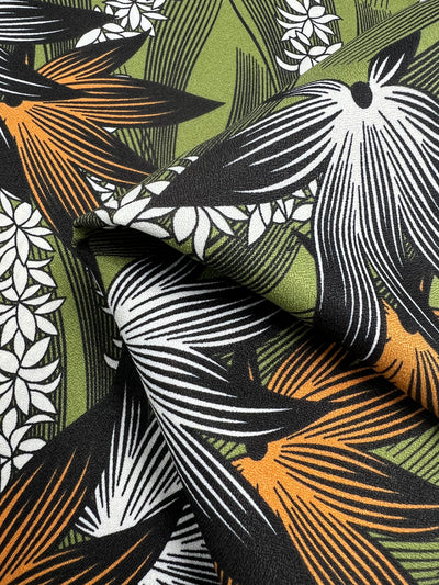 A detailed view of the lightweight Printed Crepe - Wild Hemp fabric by Super Cheap Fabrics, showcasing its striking tropical pattern with black, white, and orange flowers complemented by green leaves. The fabric is artistically folded to highlight its texture and vibrant, contrasting design elements perfect for high fashion.