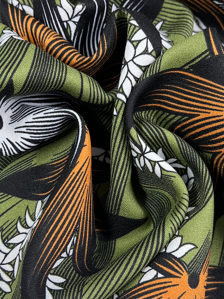 Close-up of a vibrant polyester crepe fabric called "Wild Hemp" by Super Cheap Fabrics, which is ideal for high fashion. The design includes green, orange, black, and white hues with swirling lines and petal shapes that lend a dynamic, textured look. This lightweight fabric is soft to the touch and has a slightly crumpled appearance.