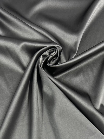 A close-up image of smooth, shiny Satin Deluxe - Pewter - 150cm fabric from Super Cheap Fabrics with a swirling fold pattern in the center, creating a spiral effect. The lightweight fabric reflects light, giving it a glossy and luxurious appearance.