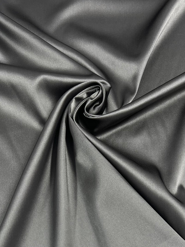 A close-up image of smooth, shiny Satin Deluxe - Pewter - 150cm fabric from Super Cheap Fabrics with a swirling fold pattern in the center, creating a spiral effect. The lightweight fabric reflects light, giving it a glossy and luxurious appearance.