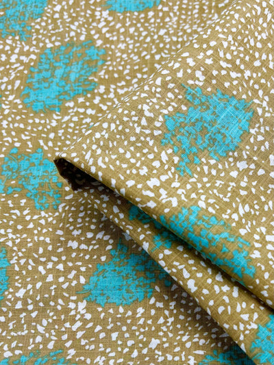 Close-up of a folded lightweight fabric featuring a pattern of small random white dots on a green background, with scattered large turquoise floral shapes. The texture of the fabric appears slightly rough, and the colors are vibrant. The fabric is "Printed Linen - Spatter - 140cm" by Super Cheap Fabrics.