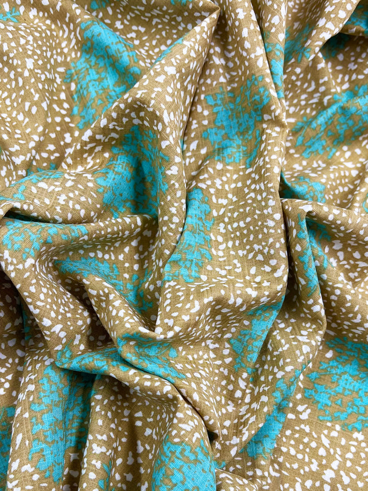 A close-up of crumpled premium fabric with an abstract design. The lightweight fabric features a pattern composed of small white dots scattered on a tan background, interspersed with irregular bright turquoise shapes, creating a dynamic, colorful texture. This is the Printed Linen - Spatter - 140cm by Super Cheap Fabrics.