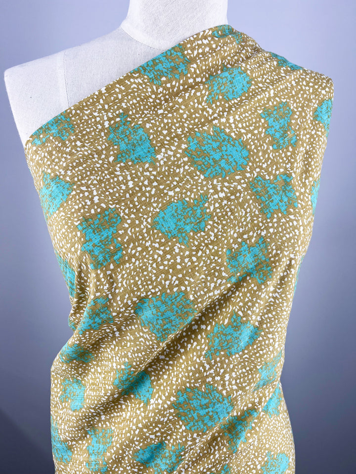 A white mannequin is draped with Super Cheap Fabrics' Printed Linen - Spatter - 140cm, featuring a brown background with scattered white specks and irregular teal blue shapes. The premium fabric is arranged across the mannequin's body diagonally from the left shoulder. The background is a gradient of light to dark gray.