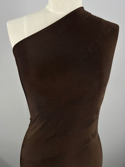 A mannequin is draped in a dark chocolate color, one-shoulder polyester fabric called Slinky Knit - Chocolate - 135cm by Super Cheap Fabrics that wraps around the torso. The smooth medium weight fabric appears to have a slight sheen, creating a subtle textured pattern under the light. The background is plain and grayish, focusing attention on the fabric and mannequin.