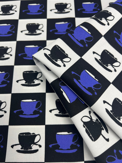 A folded piece of Super Cheap Fabrics Cotton Drill - Purple MugKnight - 155cm with a checkered pattern featuring alternating black and white squares. Each square contains an image of a blue or black teacup and saucer. The durable diagonal weave adds subtle texture, while the teacups are stylized with slight variations in illustration.