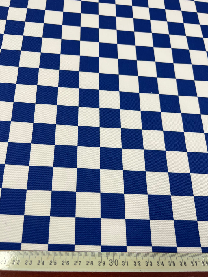 A fabric with a blue and white checkerboard pattern is shown, with each square measuring approximately 33 millimeters. A measuring tape at the bottom of the image indicates the scale, showing the Super Cheap Fabrics Cotton Drill - Blue Chess - 155cm extends beyond 30 centimeters in length.