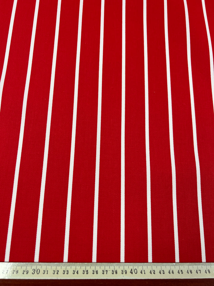 A medium-weight Denim - Bold Red - 155cm fabric by Super Cheap Fabrics with red and white vertical stripes is shown. The stripes are evenly spaced, and a wooden ruler placed horizontally at the bottom of the image indicates the measurement in centimeters, emphasizing the pattern's scale.