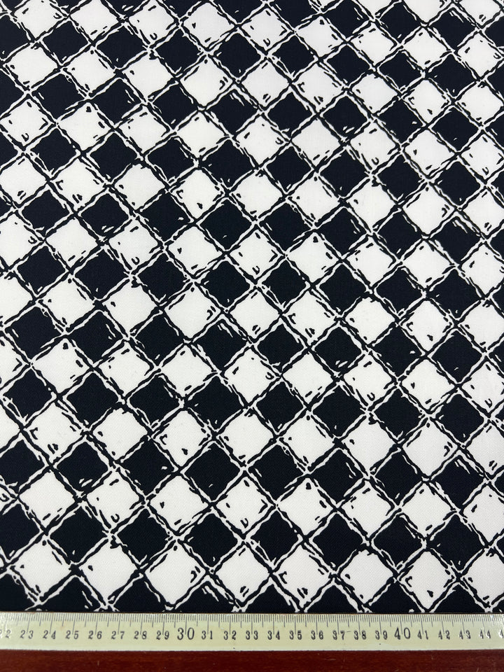 A black and white checkered fabric with an abstract pattern. The repeating square design features a rough, textured look, crafted from 100% Cotton Drill - Mosaic - 155cm by Super Cheap Fabrics. Along the bottom edge, a measuring tape displays both metric and imperial measurements.