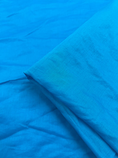 A close-up image of Cotton Voile - Vivid Blue - 135cm by Super Cheap Fabrics with a smooth texture. The extra lightweight fabric is slightly wrinkled, with a section folded over towards the right side of the image. The light highlights the texture and sheen of the 100% cotton material.