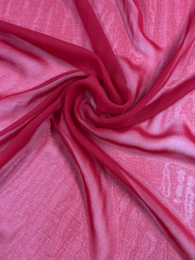 A close-up shot of sheer, red Hi-Multi Chiffon - Lava Falls - 150cm fabric from Super Cheap Fabrics loosely gathered in a swirling pattern. The lightweight fabric has a delicate, translucent texture that allows light to pass through, creating varying shades of red and pink. The swirls form a focal point in the center of the image.