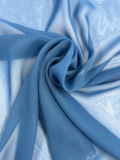 A piece of light blue, sheer fabric is shown spread out and twisted into a spiral pattern. This lightweight fabric appears delicate and translucent, with a soft and smooth texture—perfect for creating elegant floaty tops. This is the Hi-Multi Chiffon - Daphne - 150cm by Super Cheap Fabrics.