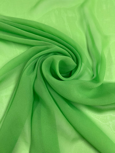 A close-up of a light green, sheer fabric with a soft, flowing texture. This 100% polyester fabric is arranged to create gentle folds and swirls, showcasing its translucence and delicate quality. The fabric featured is Hi-Multi Chiffon - Green Flash - 150cm by Super Cheap Fabrics.