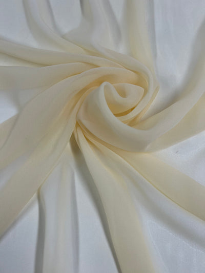 A close-up shot of a soft, sheer fabric in a pale ivory color. The 100% polyester fabric is gently twisted into a swirl shape, showcasing its delicate, thin texture and smooth appearance. The background is a plain white surface. This is the Hi-Multi Chiffon - Garden Glade - 150cm from Super Cheap Fabrics.