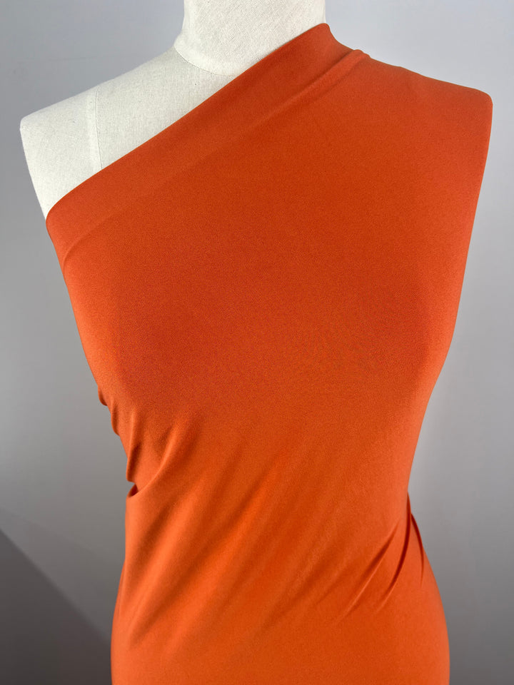 A mannequin is draped in a vibrant Super Cheap Fabrics ITY Knit - Burnt Orange - 150cm fabric. The polyester-spandex blend has a smooth texture and is wrapped tightly around the torso, highlighting the contours of the mannequin. The background is plain and gray, allowing the fabric's color to stand out.