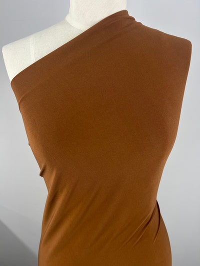 A mannequin is dressed in a one-shoulder, rust-colored fabric draped elegantly, showcasing the smooth texture and gentle folds of the ITY Knit - Rustic Brown - 150cm from Super Cheap Fabrics. The background is plain and neutral, highlighting the rich color and style of the fabric.