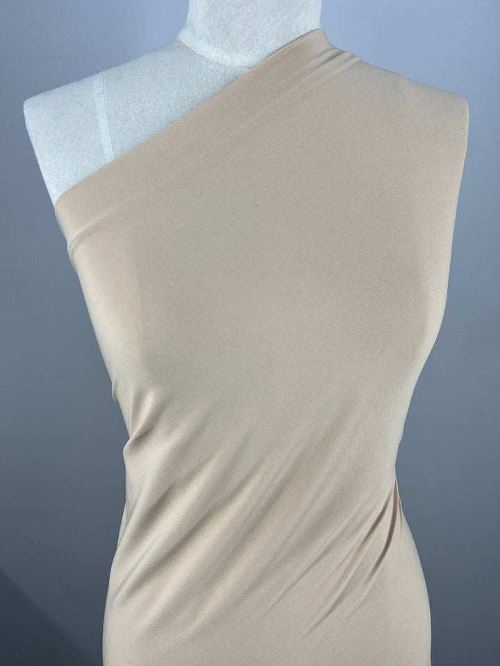 A dress form draped in a soft beige, single-shoulder fabric with smooth and flowing texture. The Super Cheap Fabrics ITY Knit - Nude - 150cm material contours the form's shape, showcasing its elasticity and elegant drape. The background is plain and unobtrusive, highlighting the opaque fabric beautifully.