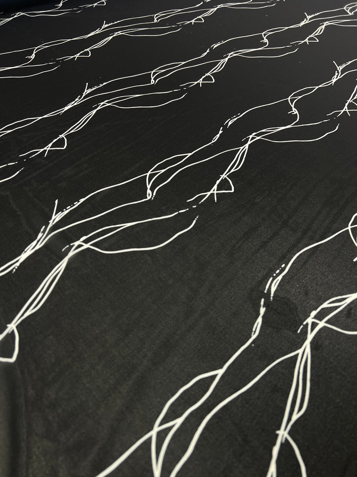A close-up of a lightweight 100% polyester surface featuring an abstract pattern of thin, white wavy lines. The lines are irregular and flow in various directions, creating a dynamic, flowing design. The contrast between the black background and white lines is striking, making it ideal for multi-use applications. This is the Designer Chiffon - Azalea - 145cm from Super Cheap Fabrics.