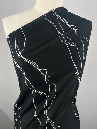A mannequin is draped in a lightweight, 100% polyester black fabric with abstract white line patterns. The material is arranged to cover one shoulder, showcasing its modern and stylish design. The plain grey background puts full focus on the pattern and texture of Designer Chiffon - Azalea - 145cm from Super Cheap Fabrics.