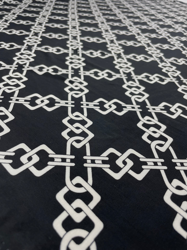 A close-up view of Super Cheap Fabrics' "Printed Lycra - Chained - 150cm" reveals a black medium weight fabric adorned with a repeating pattern of interlinked white geometric chain-like designs. The symmetrical pattern extends diagonally across this versatile fabric, creating a visually striking and intricate design.