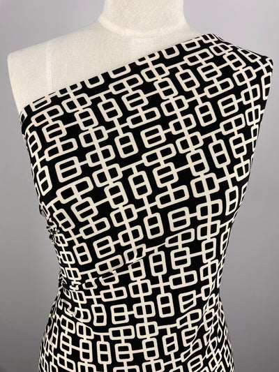 A one-shoulder dress displayed on a mannequin, featuring a black and cream geometric pattern. The medium weight polyester fabric enhances the design of interconnected rectangular shapes, creating a modern, chic look. The dress is fitted, highlighting the sleek silhouette. Made from Super Cheap Fabrics' Printed Lycra - Link - 150cm material.
