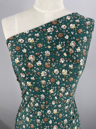 A mannequin is draped with Super Cheap Fabrics' Printed Lycra - Springy - 150cm, a medium-weight fabric that features a green base and a densely packed floral print. The polyester spandex pattern includes small white, brown, and yellow flowers along with green leaves. The dress has an asymmetrical, one-shoulder design.