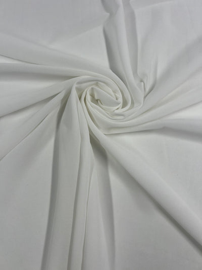 A close-up shot of Plain Georgette - Off White - 150cm from Super Cheap Fabrics with a soft, smooth texture. The fabric is arranged in a swirling pattern at the center, creating gentle folds and a sense of motion. The material appears light and airy like georgette, with subtle shadows enhancing the depth of the folds.