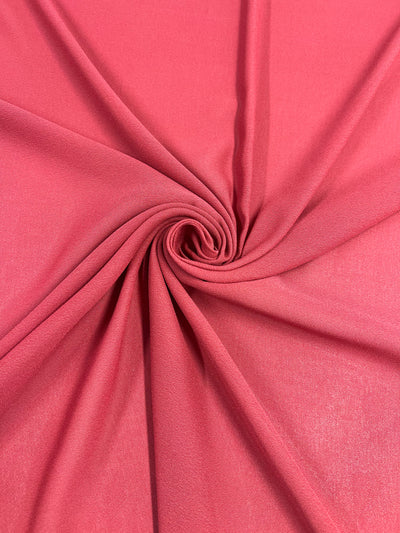 A close-up image of coral pink Plain Georgette - American Beauty - 150cm by Super Cheap Fabrics, with the material gathered and twisted at the center, creating a spiral pattern. The lightweight fabric appears smooth and slightly shiny, with folds radiating outward from the center twist.