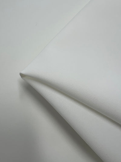 A close-up photo captures a neatly folded piece of Stretch Suiting - Ivory - 150cm by Super Cheap Fabrics, angled perfectly against a white background.
