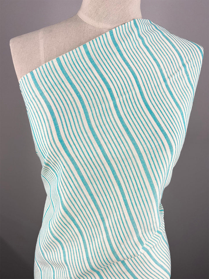 A mannequin is dressed in a one-shoulder, asymmetrical garment made of 100% Cotton Voile - Light Blue- 150cm by Super Cheap Fabrics with irregular, wavy, teal stripes. The lightweight fabric drapes elegantly across the torso, showcasing the striped pattern with a silky finish. The background is plain gray.