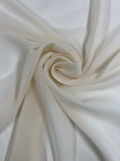 A close-up of a beige and white lightweight fabric swirled in a circular pattern. The Hi-Multi Chiffon - Nude - 150cm from Super Cheap Fabrics appears soft and sheer, with a smooth and flowing texture. The gentle folds and subtle color gradient create a delicate and elegant appearance, perfect for floaty tops.
