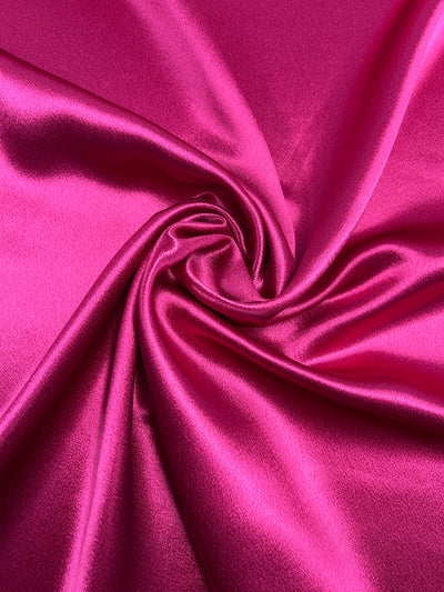 Close-up image of vibrant fuchsia fabric with a glossy sheen. The lightweight **Satin Back Crepe - Fuchsia - 112cm** from **Super Cheap Fabrics** appears smooth, and its folds are arranged in a spiral pattern, creating a dynamic and elegant appearance. The lighting emphasizes the fabric's silky texture and lustrous finish.