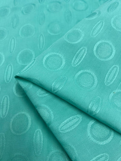A lightweight fabric in a stunning teal color with an embossed circular pattern is shown. The circles vary slightly in shading, giving a textured appearance overall. Made from soft rayon fabric, it is folded to showcase the intricate design from different angles. Introducing the Super Cheap Fabrics Textured Rayon - Ice Green - 140cm.