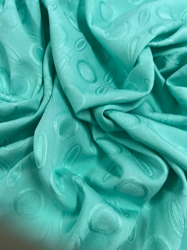 A close-up of a crumpled, silky Textured Rayon - Ice Green - 140cm from Super Cheap Fabrics in a vibrant ice green color. The light weight fabric features an embossed pattern of variously sized ovals and circular shapes, adding texture and depth to the material.