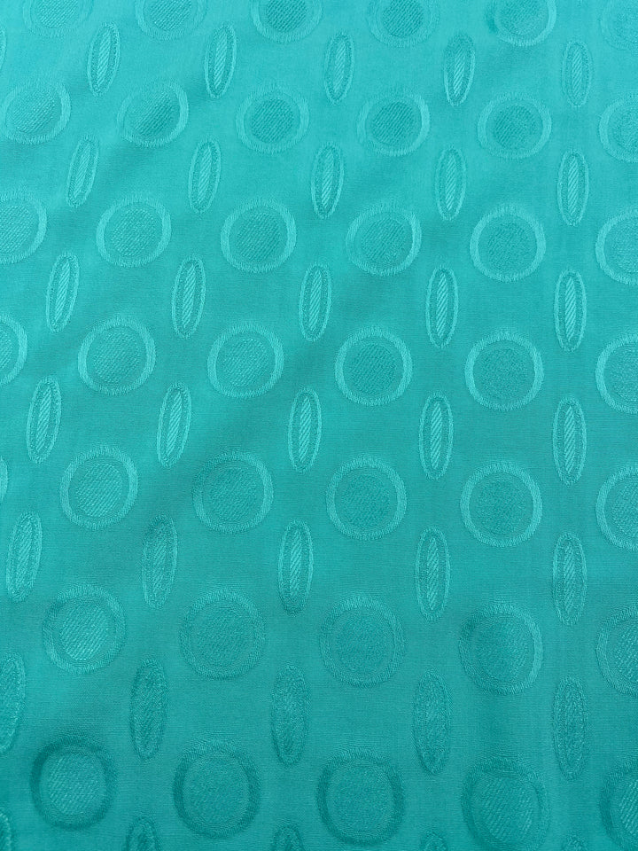 A turquoise Textured Rayon - Ice Green - 140cm by Super Cheap Fabrics with a repeating pattern of embossed ovals and circles, arranged in a grid-like formation. The texture is slightly raised, giving the light weight fabric a subtle, elegant design. The ice green background has a smooth, silky appearance.