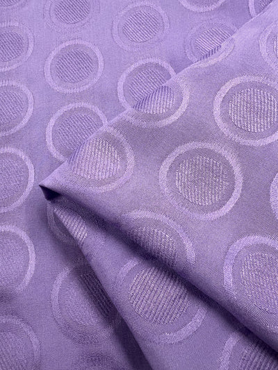 A close-up image of folded lavender Textured Rayon - Fairy Wren - 140cm fabric from Super Cheap Fabrics with a textured pattern of concentric circles. The surface has a subtle sheen, and the circular designs are evenly spaced, adding a sense of rhythm to the fabric's silky appearance, making it perfect for lightweight swing tops and dresses.