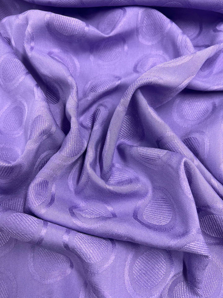 A close-up of crumpled lavender fabric featuring a textured circular pattern. The Super Cheap Fabrics Textured Rayon - Fairy Wren - 140cm has a soft sheen, creating subtle variations in color and texture as it catches the light. The circular design adds a decorative touch to the smooth, silky material, perfect for swing tops and dresses.