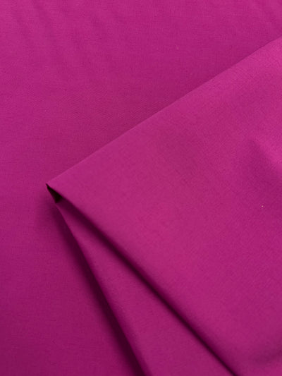 A piece of vibrant Poplin - Cactus Flower - 152cm from Super Cheap Fabrics is neatly folded on one corner. The material appears smooth and slightly glossy, showcasing a rich, deep color with a fine texture. This lightweight fabric is photographed against a background of the same hue.